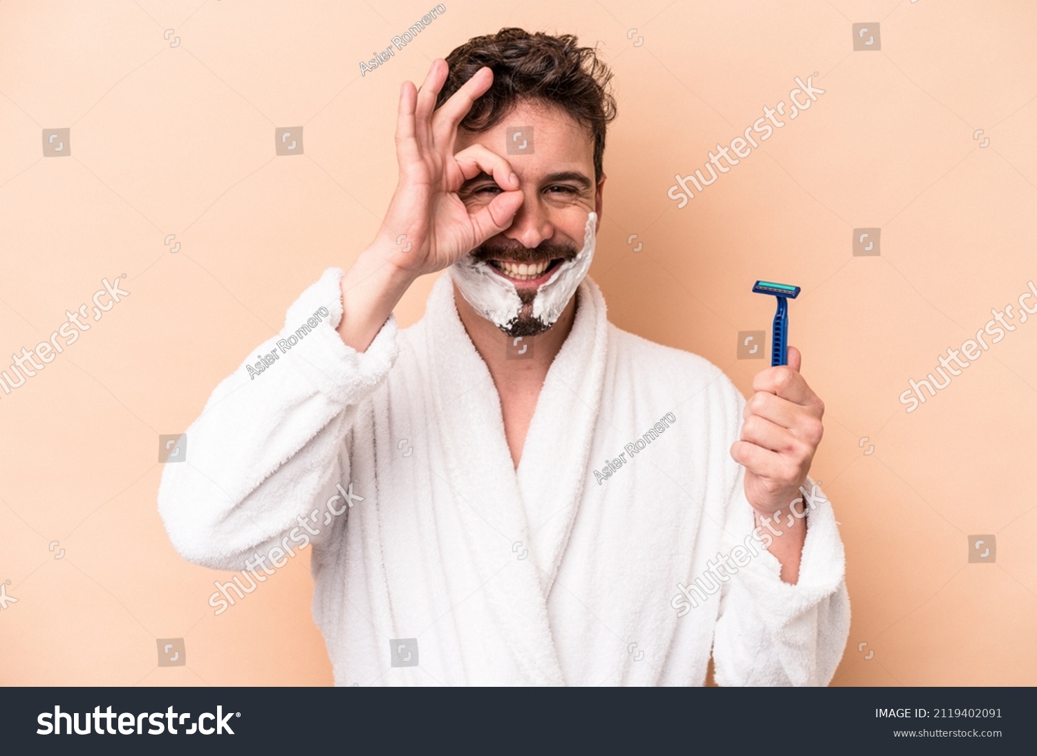 Young caucasian man wearing shaving foam and holding razor blade isolated on beige background excited keeping ok gesture on eye. #2119402091