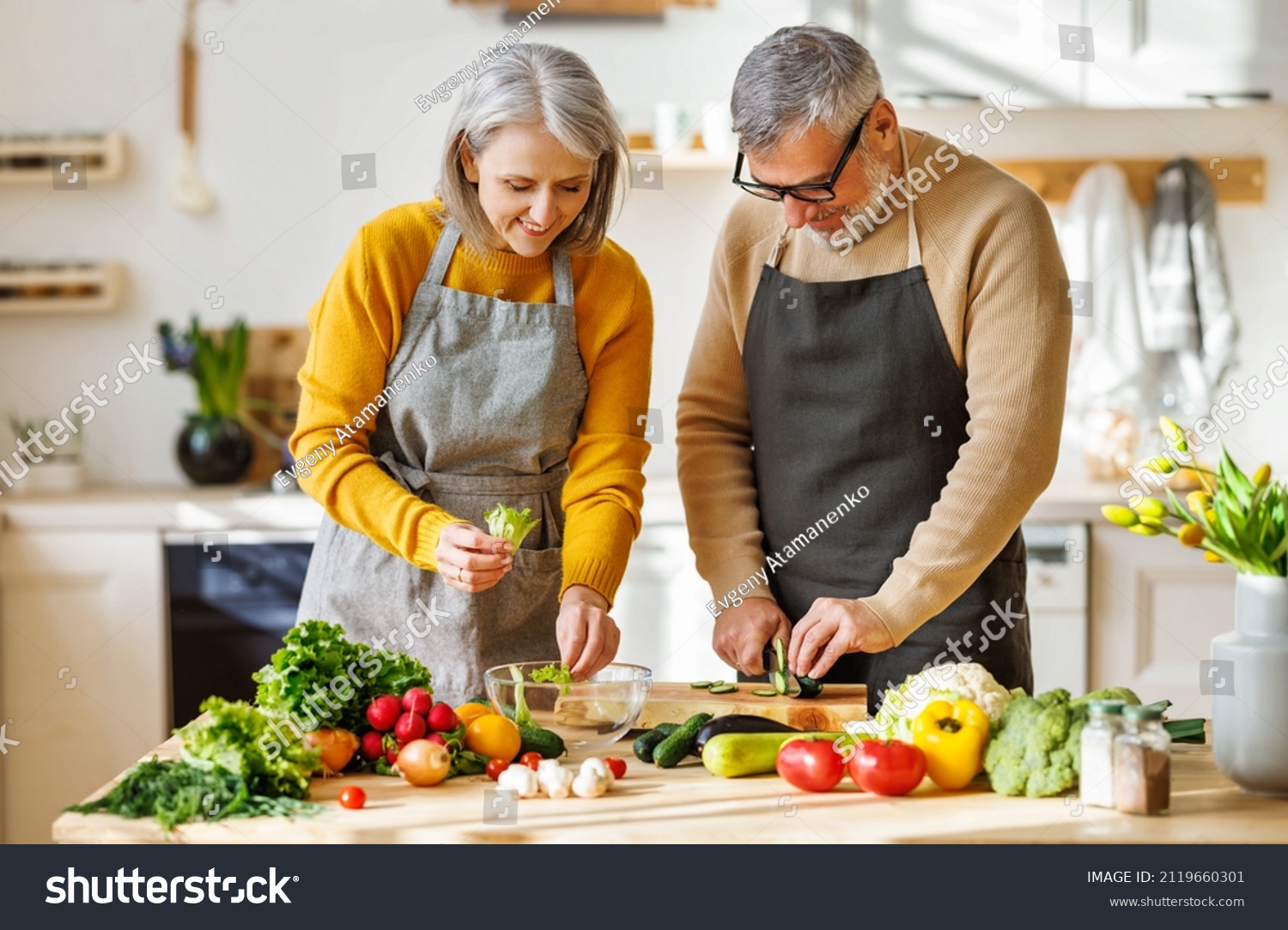 Happy elderly couple smiling husband and wife in aprons prepare salad together at kitchen table, chopping variety of colorful vegetables, trying to maintain healthy lifestyle eating vegetarian food #2119660301