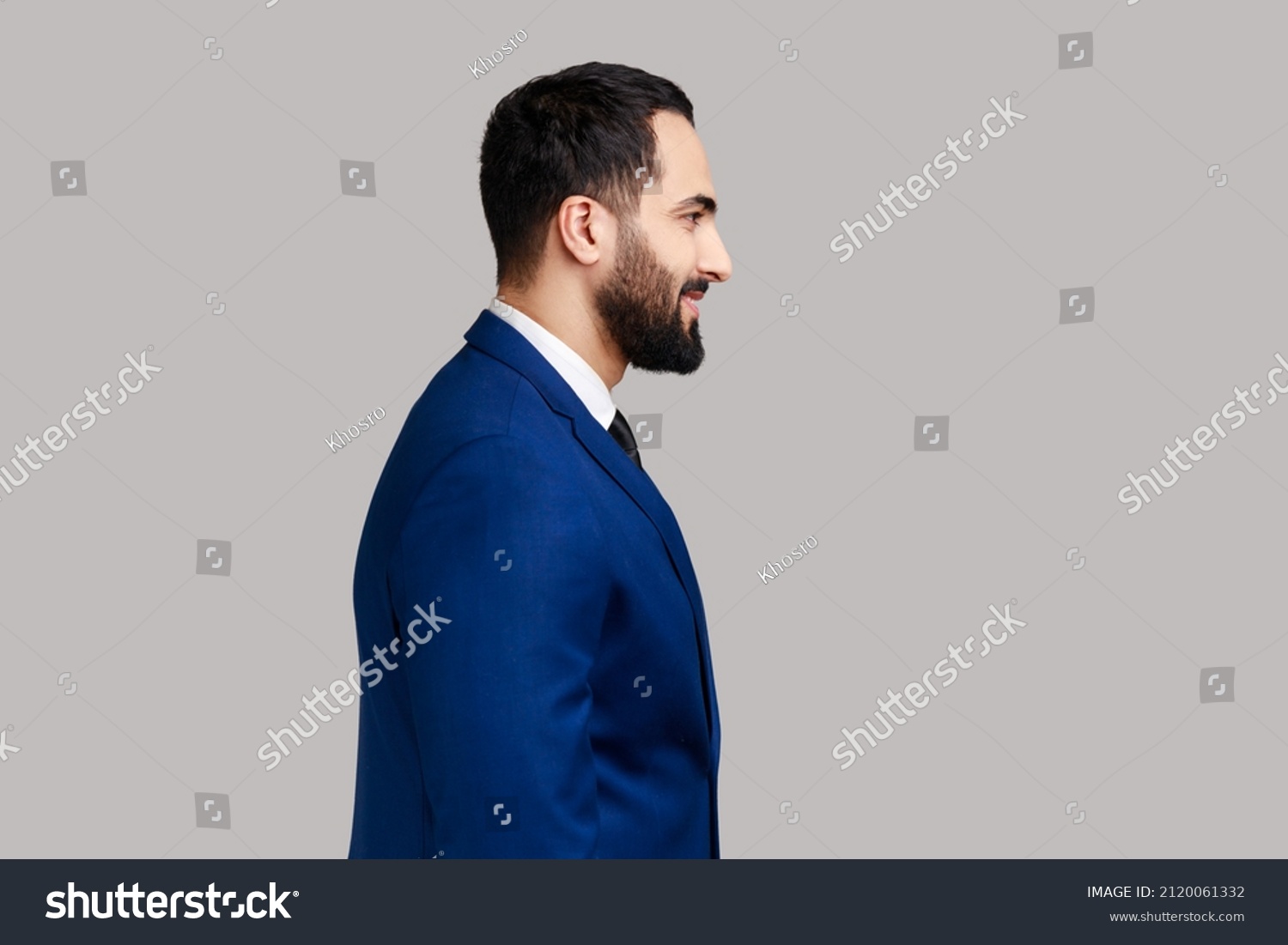 Side view portrait of cheerful bearded man with smile, standing and looking at camera, expressing positive emotions, wearing official style suit. Indoor studio shot isolated on gray background. #2120061332