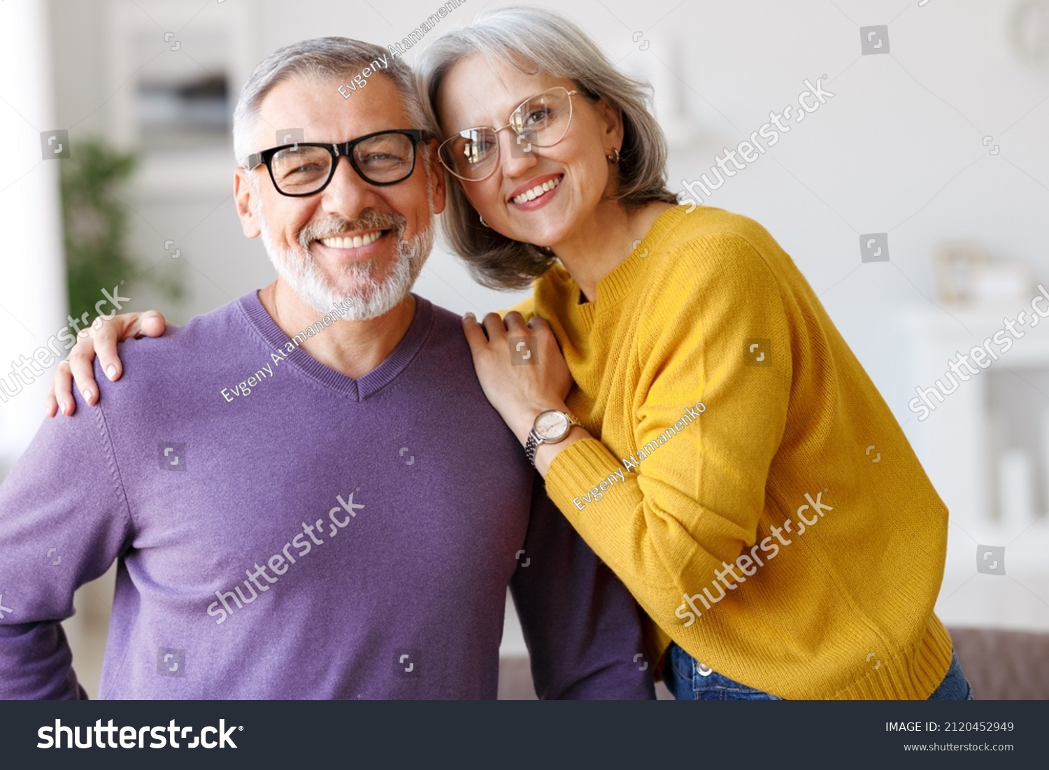 Portrait of happy beautiful senior caucasian family couple in love smiling at camera, retired man and woman hugging embracing while relaxing on sofa in living room at home, enjoying life on retirement #2120452949