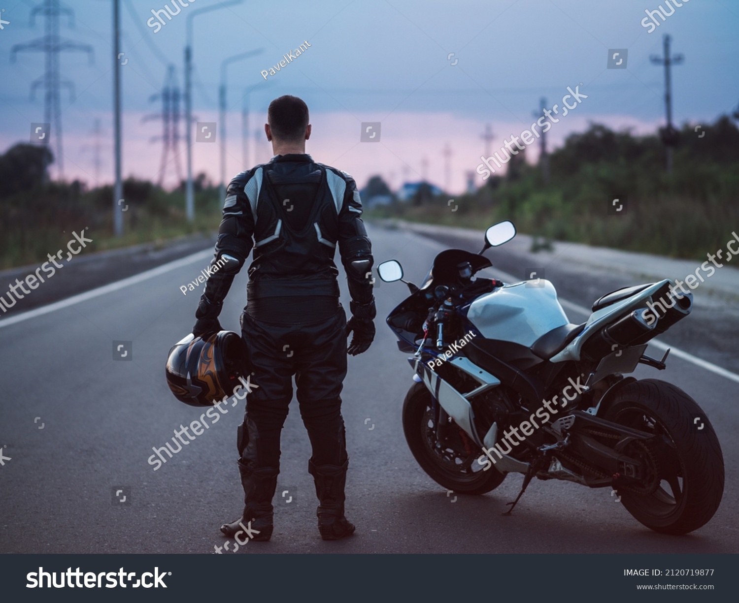A biker stands next to a motorcycle on an asphalt road and looks into the distance. Summer sunset #2120719877