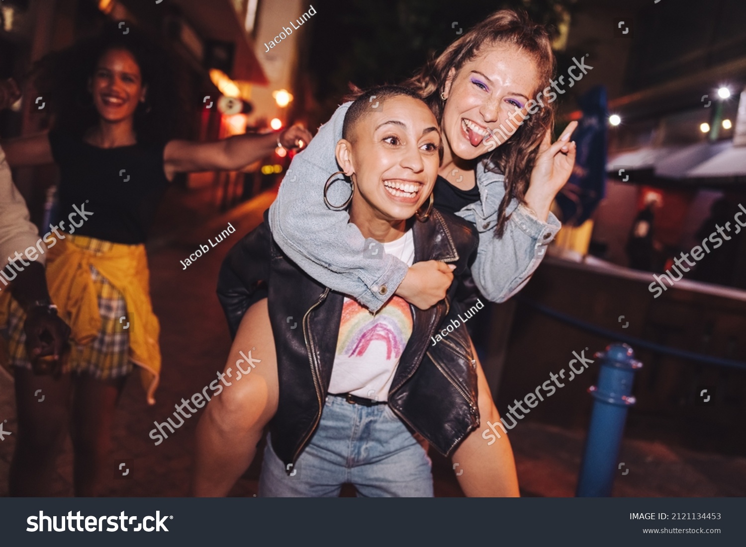 Playful young woman piggybacking her best friend outdoors at night. Happy young women having fun while going out with their friends in the city. Vibrant friends spending their weekend together. #2121134453