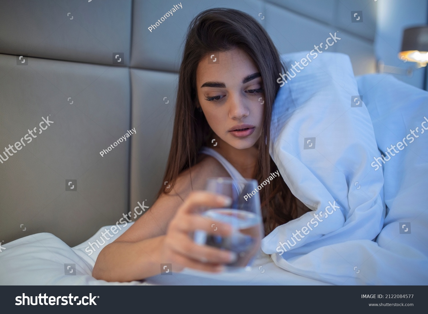 Young woman drinking glass of water in bed at night. Woman drinking a glass of water before going to sleep, she is lying in bed #2122084577