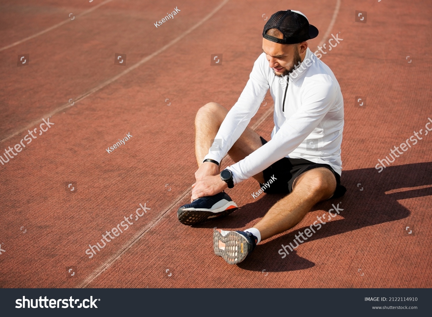 male runner in white black clothes holds his knee at the stadium, sports injury #2122114910