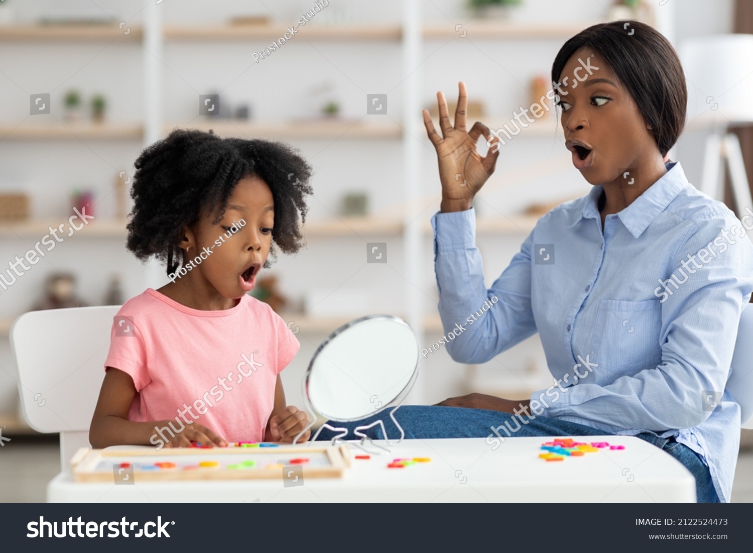 Pretty little black girl attending speech therapy session at clinic, looking at mirror, beautiful young african american woman speech-language pathologist working on sounds with kid, articulating #2122524473