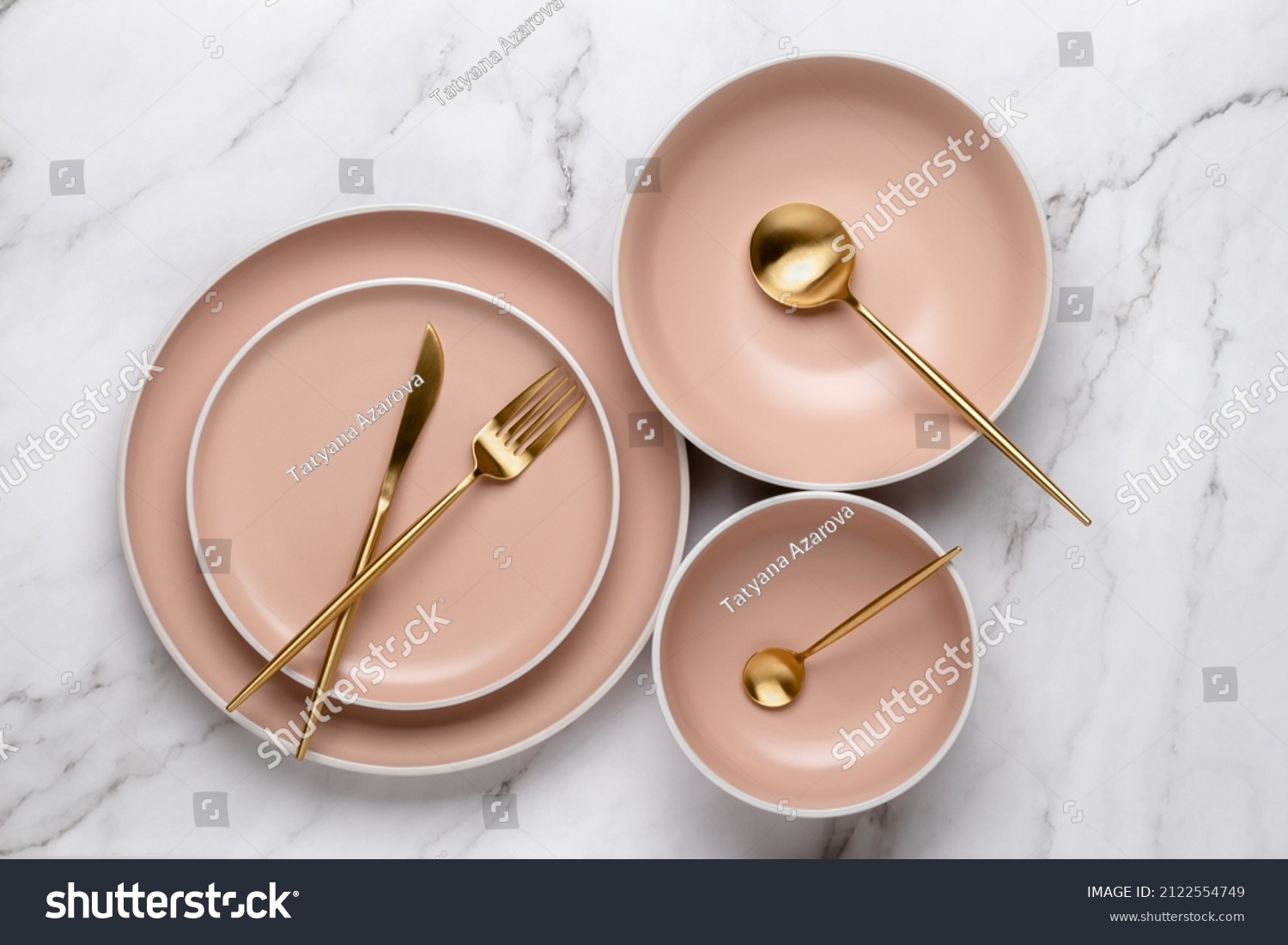 Dishes and utensils for serving and eating meals. Beige round rimmed plates and gold colored cutlery on a white marble table, top view. Modern ceramic crockery, trendy tableware #2122554749