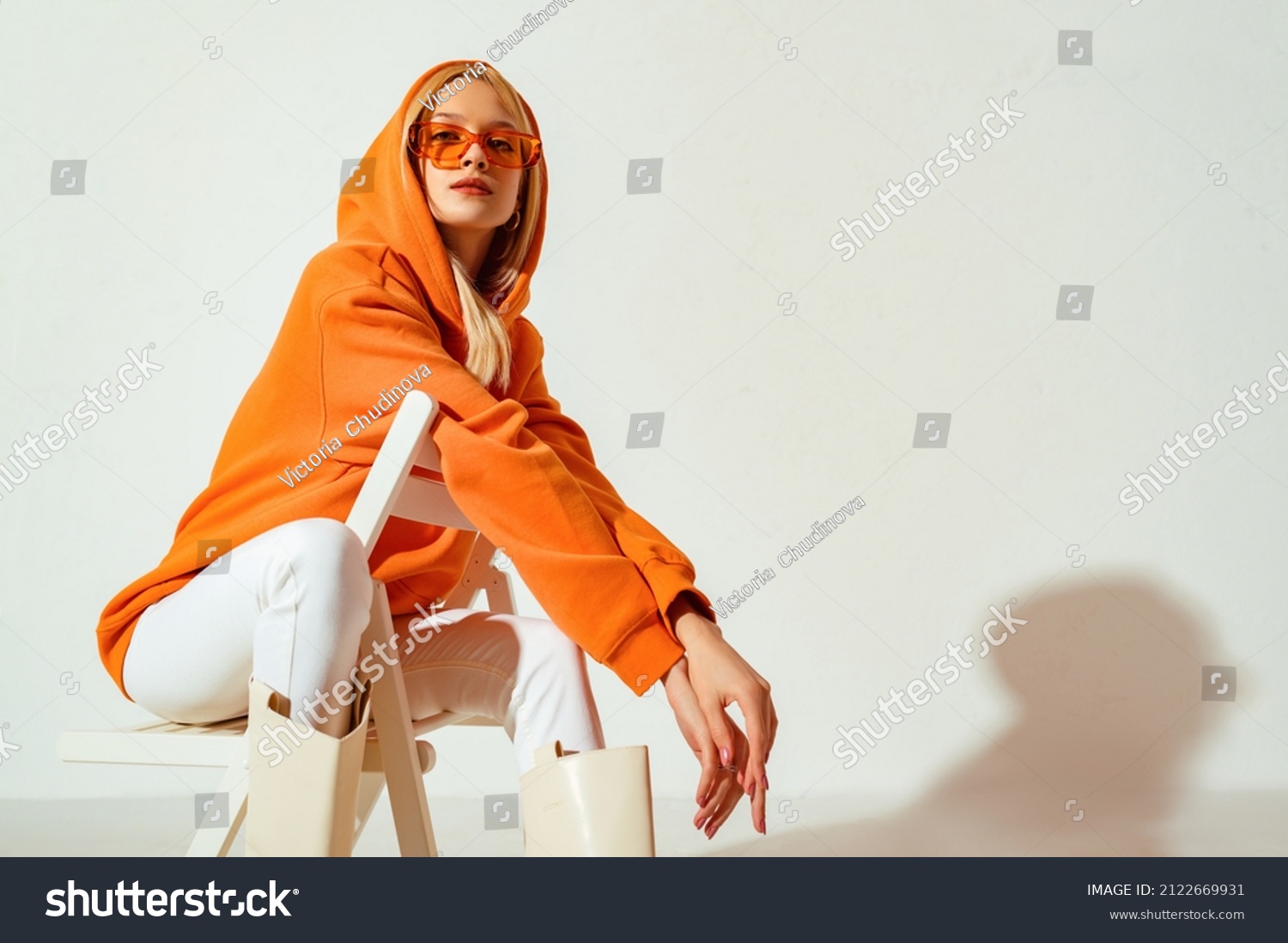 Young confident blonde girl wearing trendy orange hoodie, color sunglasses, posing on white background. Studio fashion portrait. Copy, empty space for text #2122669931