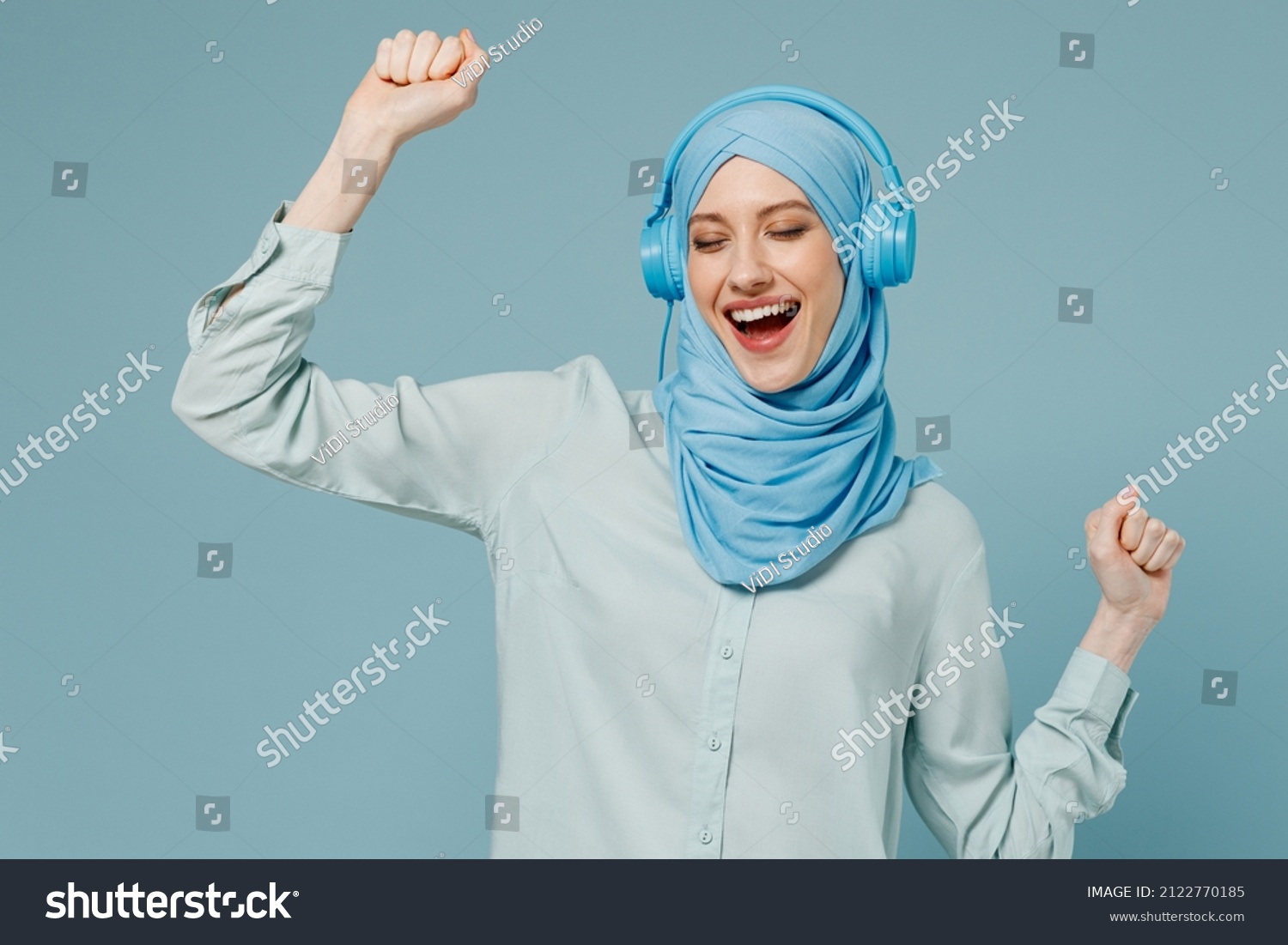Young smiling arabian asian muslim woman in abaya hijab headphones listen to music dancing have fun isolated on plain blue background studio portrait. People uae middle eastern islam religious concept #2122770185