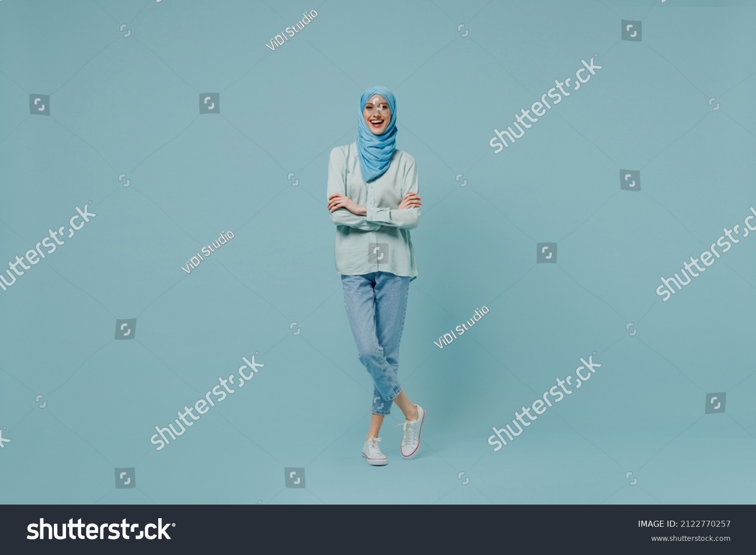 Full body young arabian asian muslim woman in abaya hijab hold hands crossed folded look camera isolated on plain blue background studio portrait. People uae middle eastern islam religious concept. #2122770257