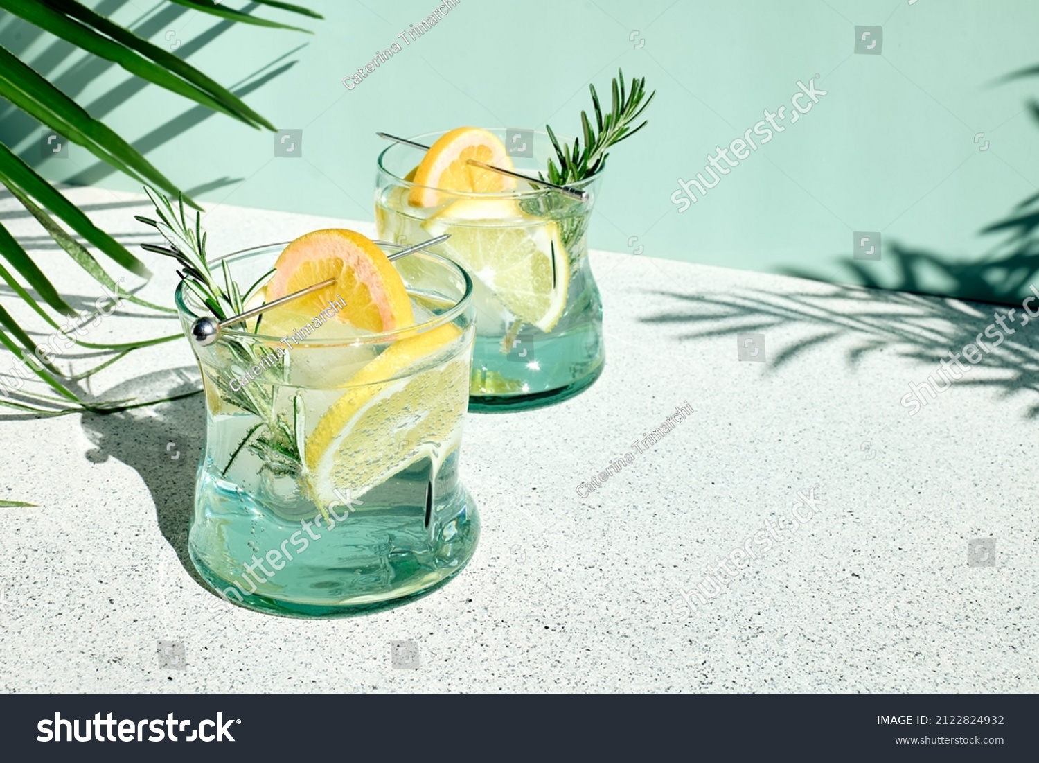 Summer refreshing lemonade drink or alcoholic cocktail with ice, rosemary and lemon slices on pastel light green surface. Fresh healthy cold lemon beverage. Water with lemon. #2122824932