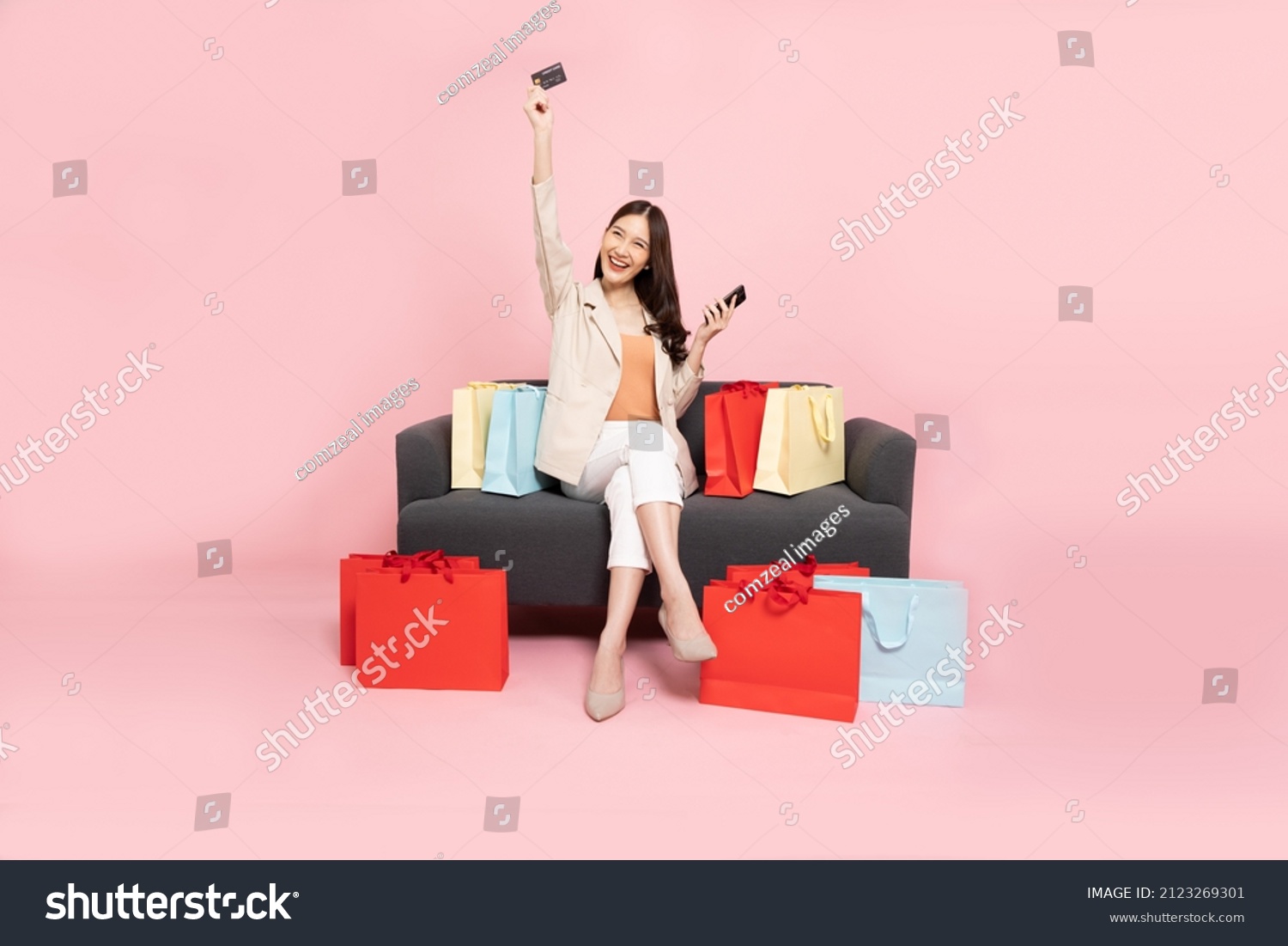 Full length portrait of Excited Asian woman sitting on sofa with shopping bags and hands up with credit card and mobile phone isolated on pink background, Shopper or shopaholic concept #2123269301