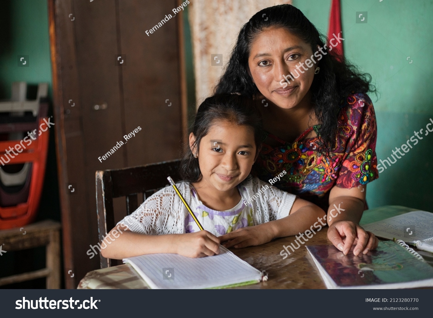 Hispanic mom helping her little daughter do her homework - Mom teaching her daughter to read and write at home - Mayan family at home #2123280770