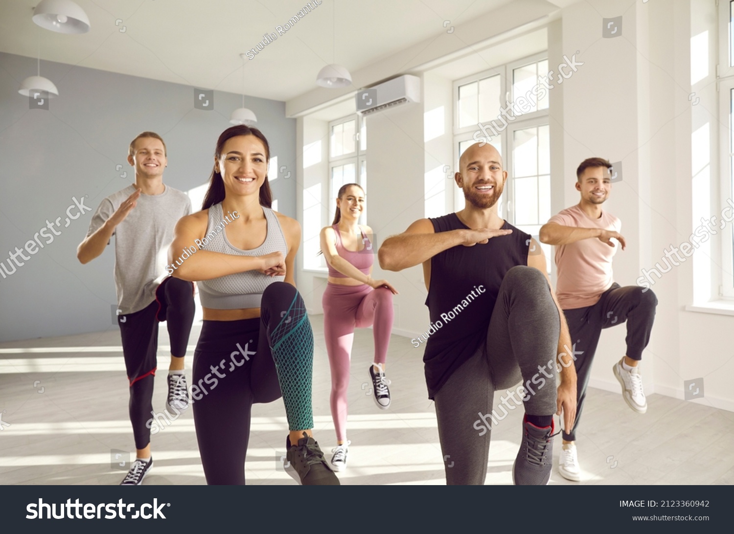 Group of young sporty people doing exercises together in bright and spacious gym. Happy men and women doing aerobics exercises lifting high knees in gym. Fitness, sport, aerobics and people concept. #2123360942