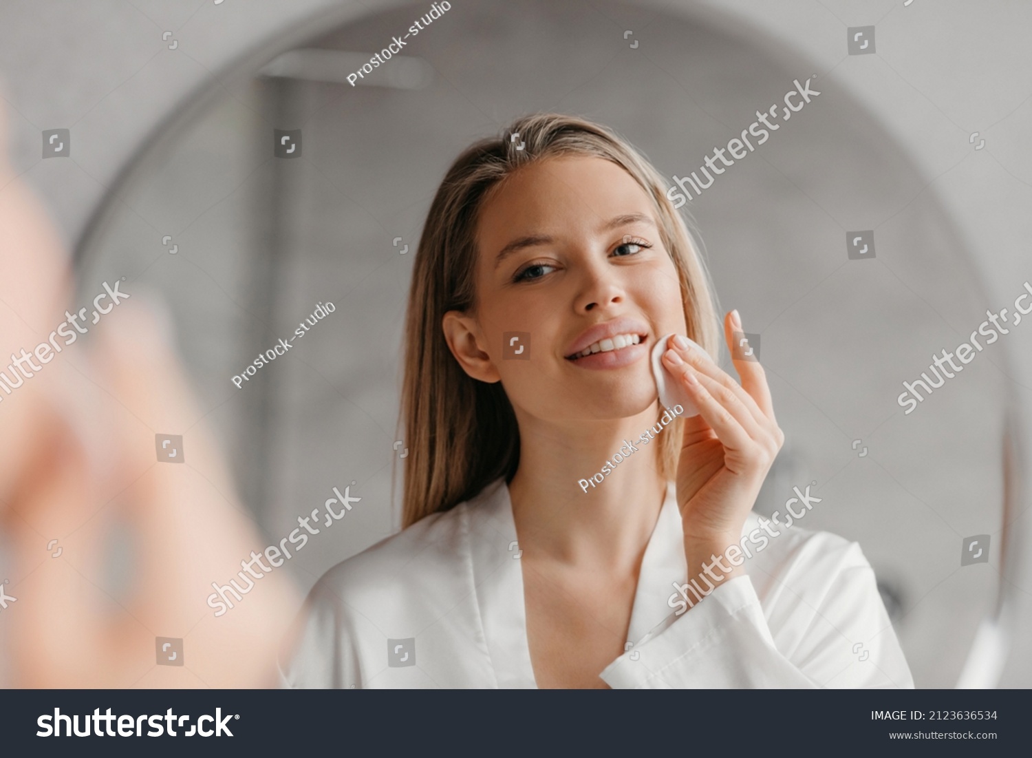 Young woman caring for facial skin using cotton pad and looking in round mirror in bathroom interior. Beauty care and pampering. Daily female skincare routine concept #2123636534
