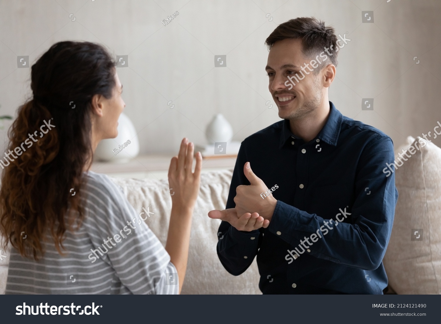 Smiling man and woman with deafness using signs for communication, sitting on couch at home, smiling. Therapist teaching gesture language to patient with disability. Hearing disorder concept #2124121490