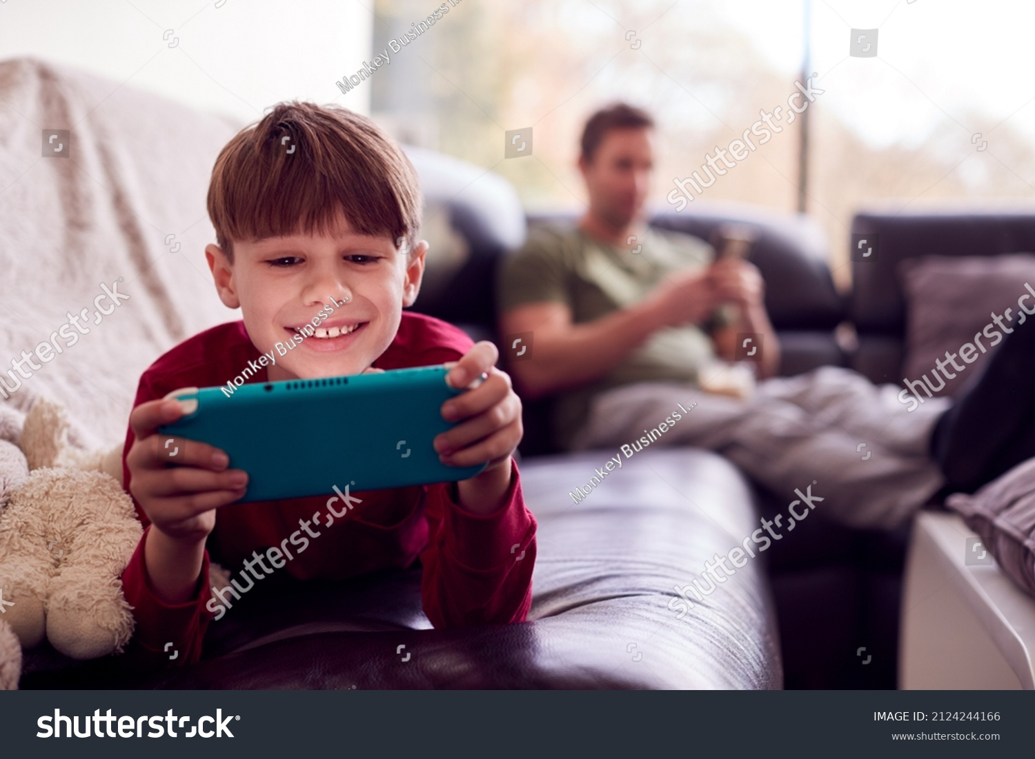 Father Uses Mobile Phone As Son Plays Computer Game On Portable Gaming Device At Home In Pyjamas #2124244166
