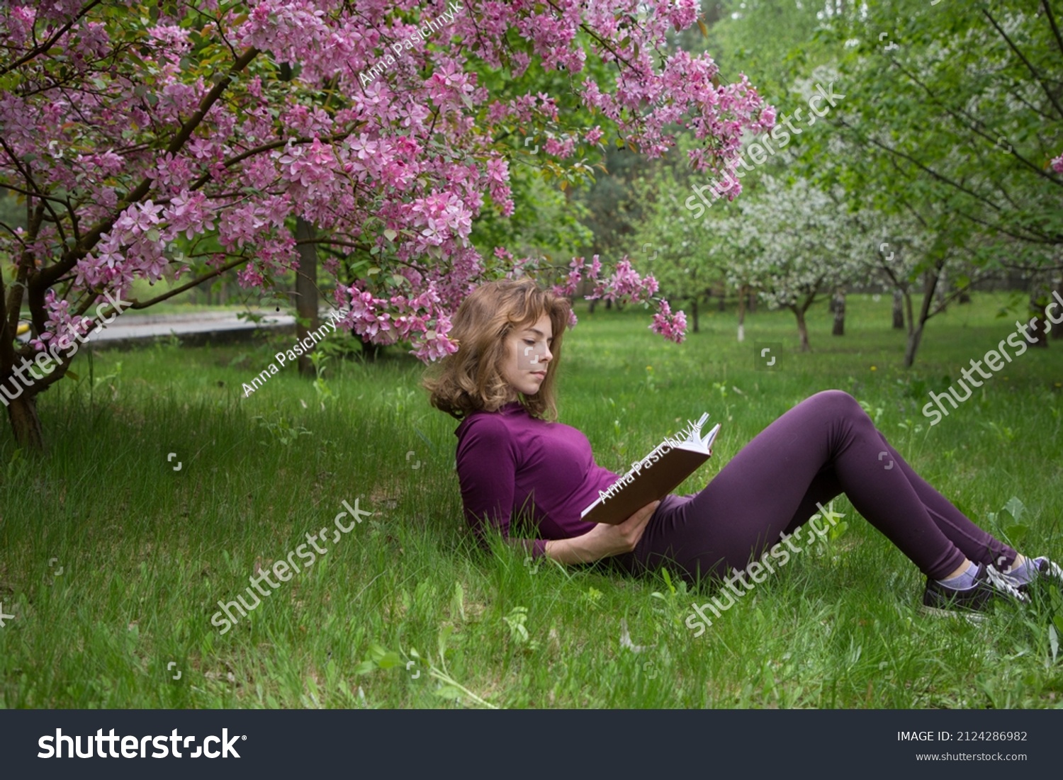 young woman, student, reads book with interest, lying under tree on spring day in park. girls - teenager 17 years old with book on grass. preparation for exams. Spring leisure. Passion for reading #2124286982