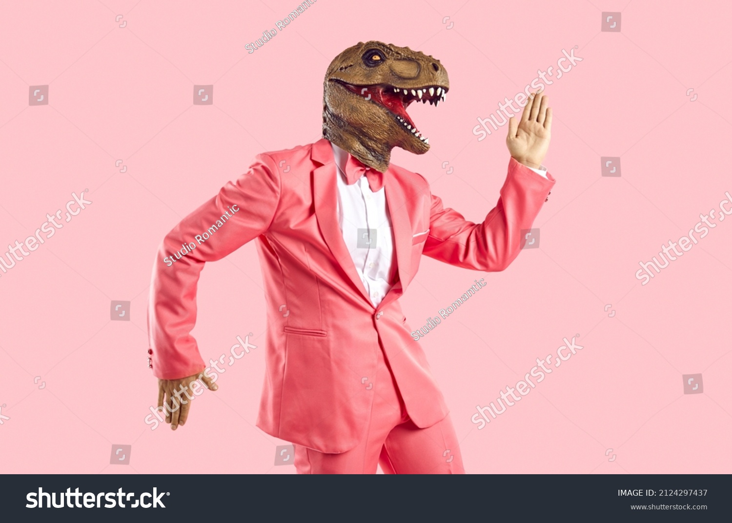 Funny man in rubber dinosaur mask dancing and having fun in the studio. Happy lizard headed guy in stylish funky vibrant pink party suit doing Egyptian dance moves isolated on pink colour background #2124297437
