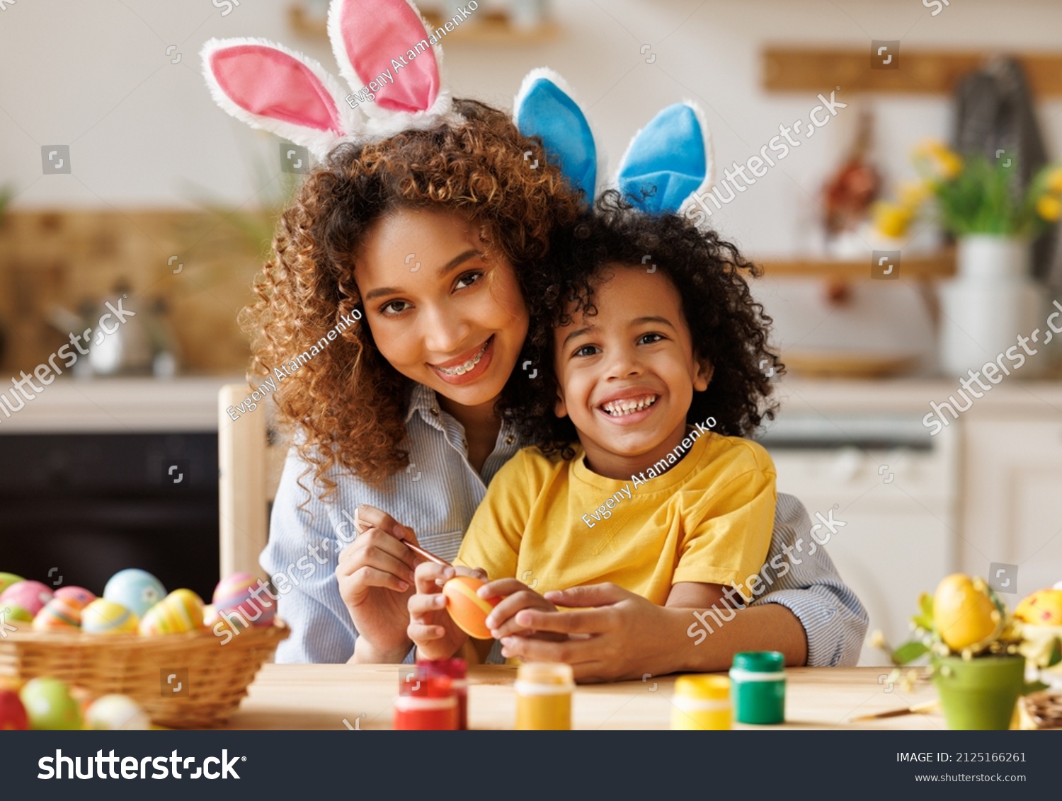Easter Family traditions. Loving ethnic young mother teaching happy little kid soon to dye and decorate eggs with paints for Easter holidays while sitting together at kitchen table  #2125166261