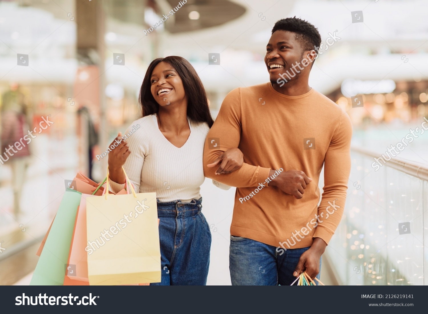 Portrait of beautiful millennial African American guy and lady shopping on weekend, walking in the city mall with colorful bags, looking at boutique window and laughing spending time together #2126219141