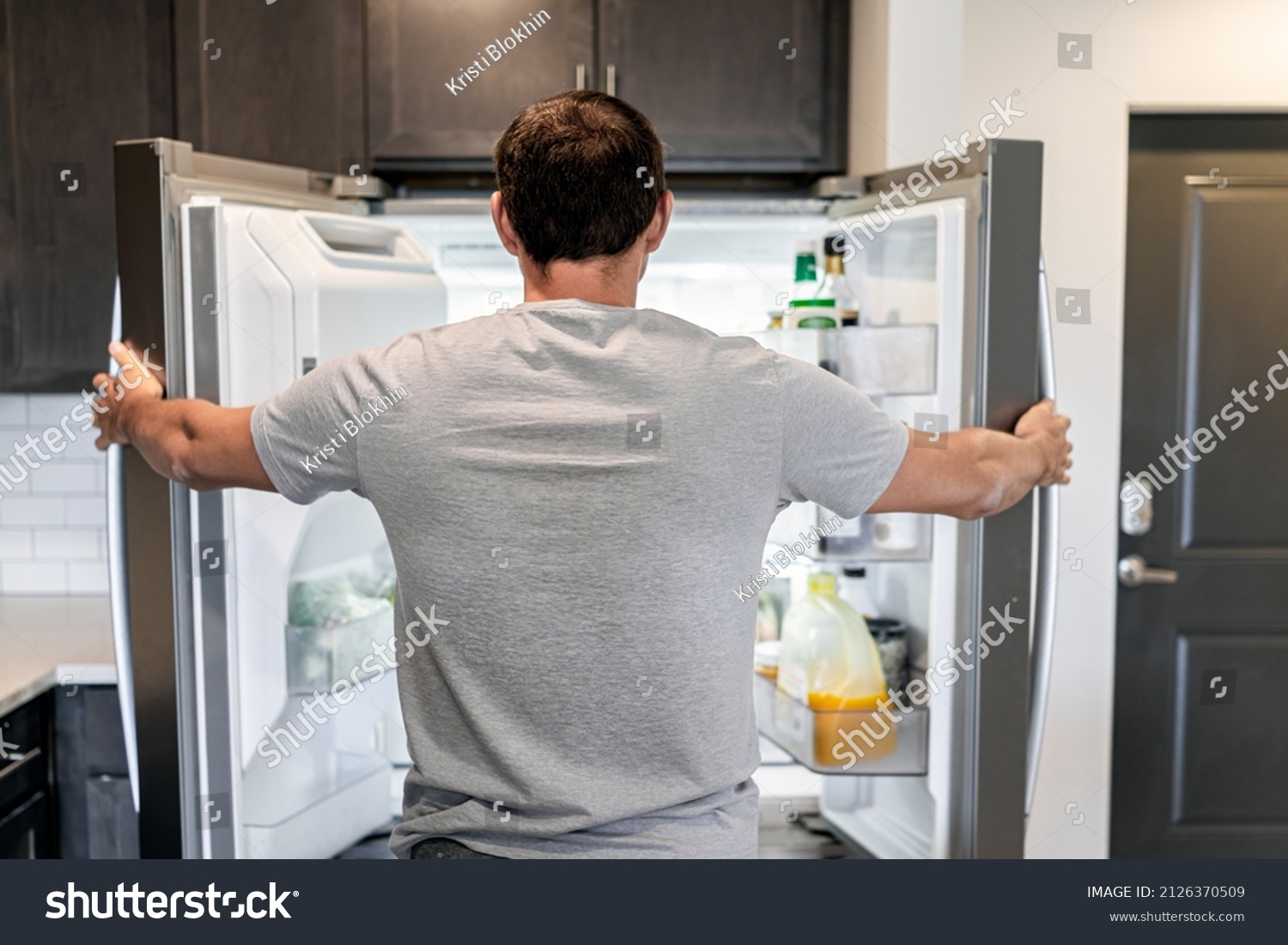 Back of hungry man opening fridge refrigerator doors domestic appliance searching for food inside with condiments and juice in modern kitchen #2126370509