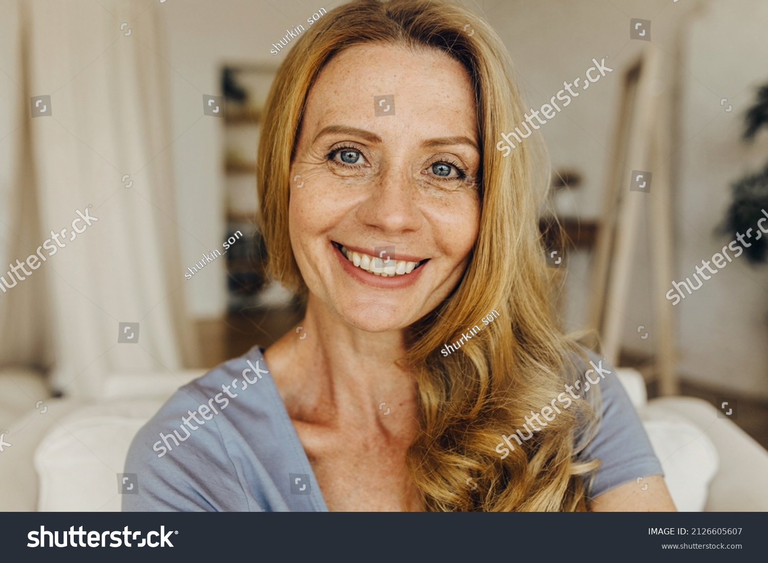 Senior woman-blogger with long golden hair, pretty freckled face, blue eyes and charming smile making selfie or recording stories for social network page, looking friendly and happy #2126605607