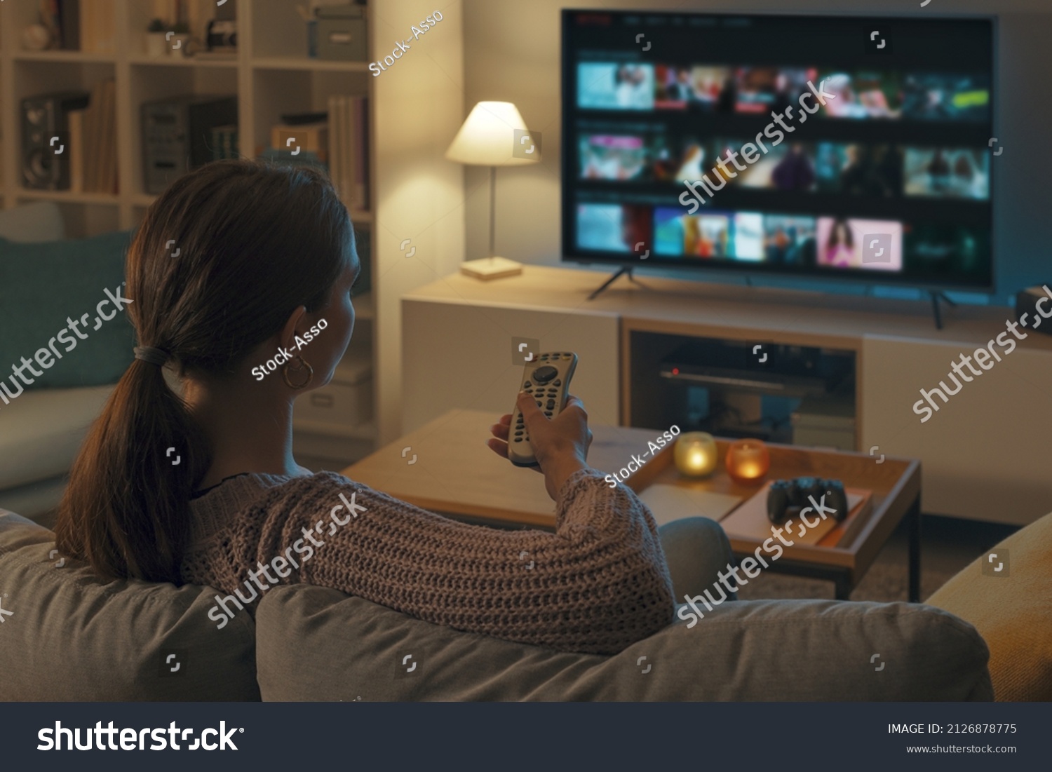 Woman relaxing on the couch, she is using the remote control and choosing a TV show or movie on the television menu #2126878775