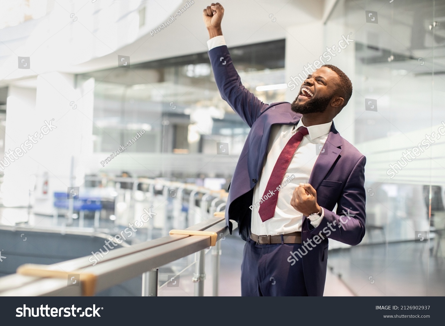 Cheerful african american young businessman celebrating success, handsome black manager in suit and tie raising hands up and screaming, office building interior, panorama with copy space #2126902937