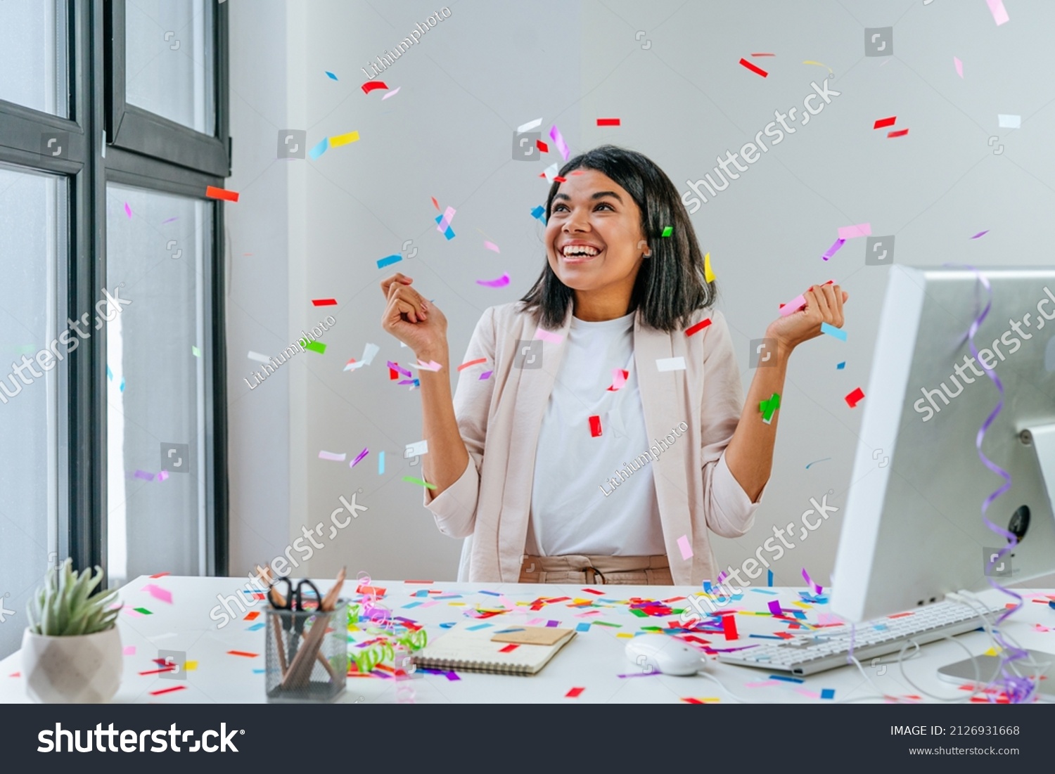 Young business woman having fun time catching confetti sitting at the desk in the office. Party time on the work place. Selective focus. #2126931668