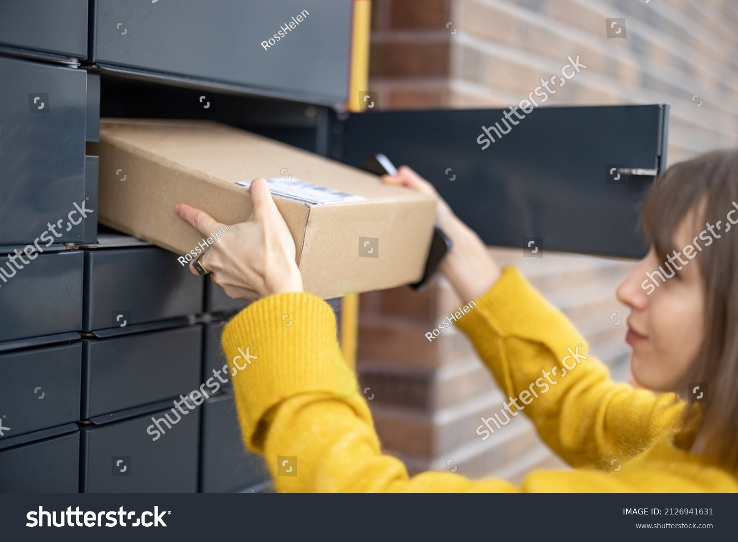 Young woman getting parcel from cell of automatic post terminal outdoors. Concept of contactless and smart delivery. Idea of modern shipping and logistics. Woman wearing yellow sweater #2126941631