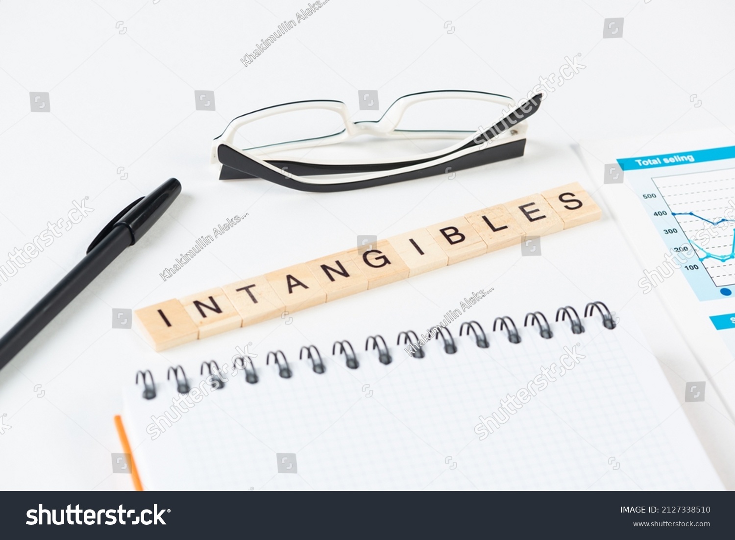 Intangibles concept with letters on wooden cubes. Still life of office workplace with supplies. Flat lay white surface with glasses, pen and notepad. Intellectual capital and property. #2127338510
