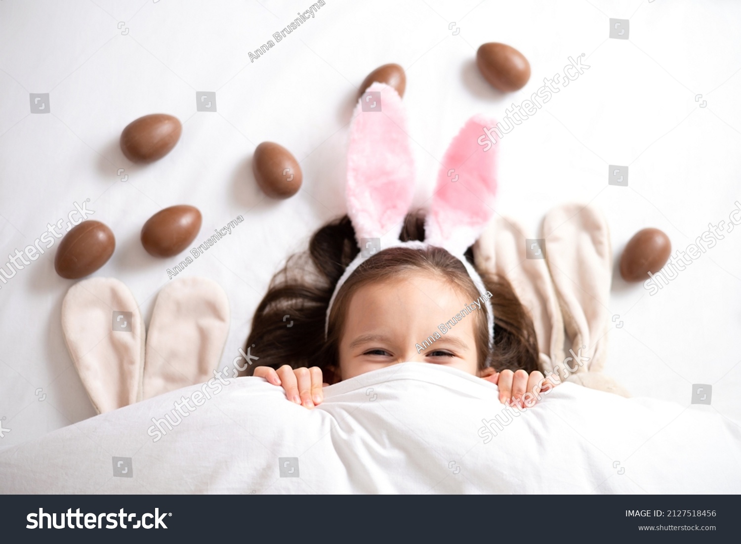 Happy Easter. cute beautiful girl with blue eyes and bunny ears peeking out from under the blanket. Chocolate eggs are scattered on the bed. Egg hunt. Lifestyle High quality photo #2127518456