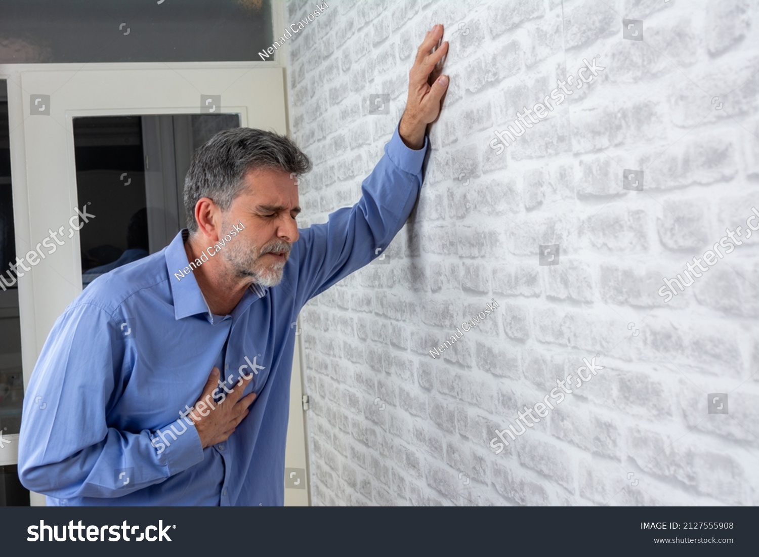 Senior man presses hand to chest has heart attack suffers from unbearable pain closes eyes wears  poses against grey brick wall background. People age and problems with health concept, copy space. #2127555908
