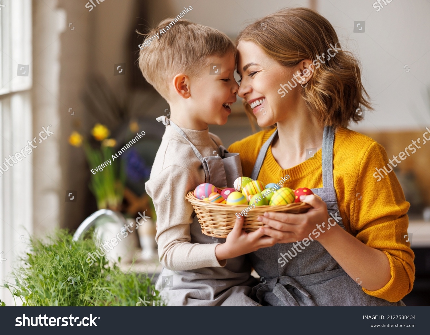 Sweet family portrait of happy mother and little son holding wicker basket full of painted multi-colored Easter eggs, tenderly embracing and smiling in cozy light kitchen at home, selective focus #2127588434