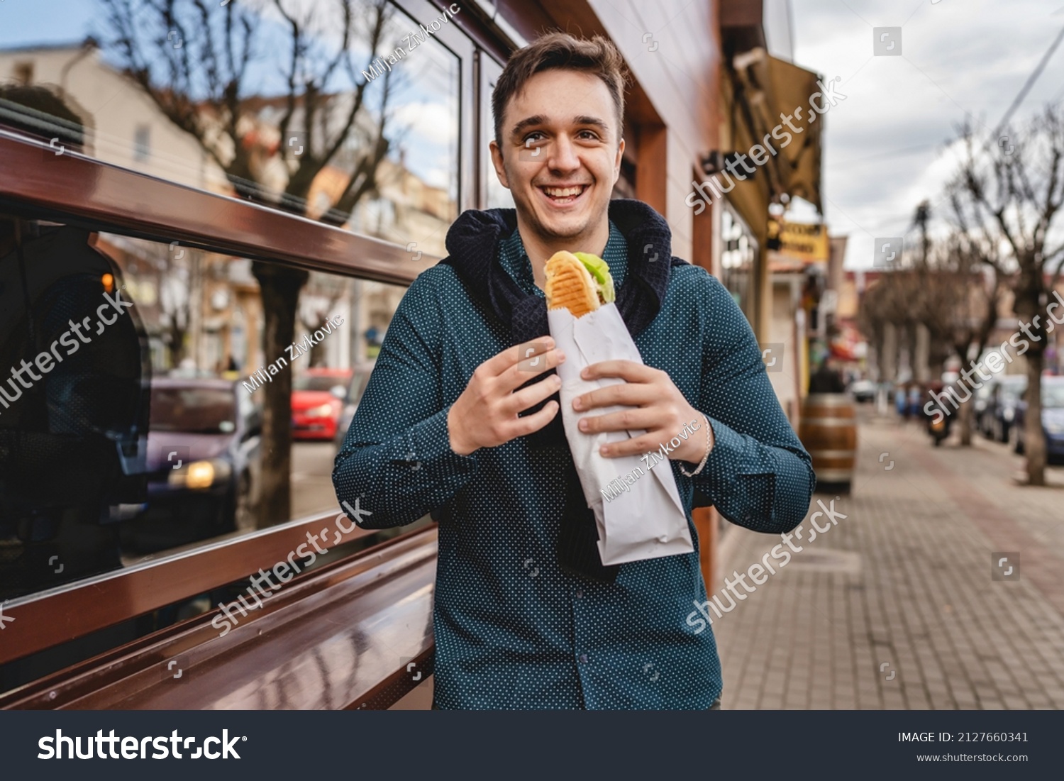 One man standing on the street in autumn spring or winter day holding sandwich wearing jacket eating on the feet while waiting outdoor in front of building real people copy space fast food concept #2127660341