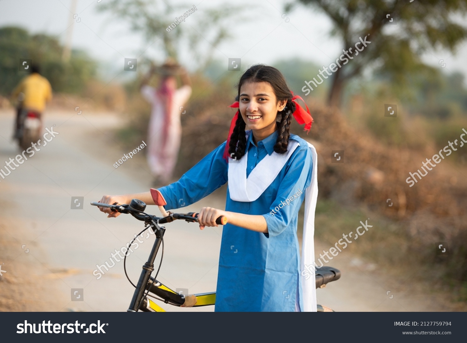Portrait of happy young rural indian girl wearing blue school uniform standing with bicycle at village street. #2127759794
