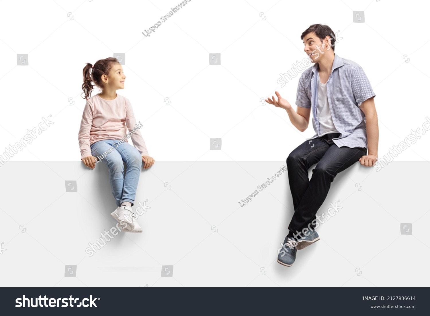 Young man sitting on a blank panel and talking to a child isolated on white background #2127936614