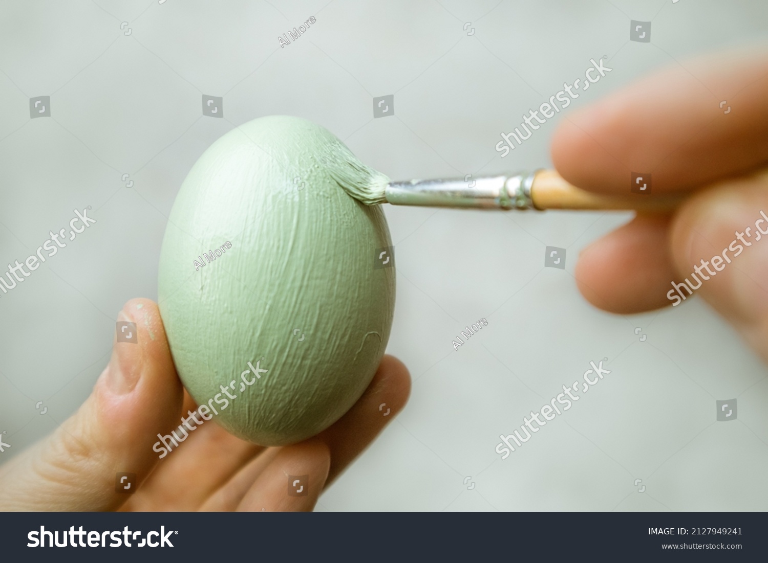 Hand holds brash and paints egg. A process of painting an Easter egg with light green paint. Paint brush in woman's hand on gray background. Tradition and people concept. Spring season, April holiday. #2127949241