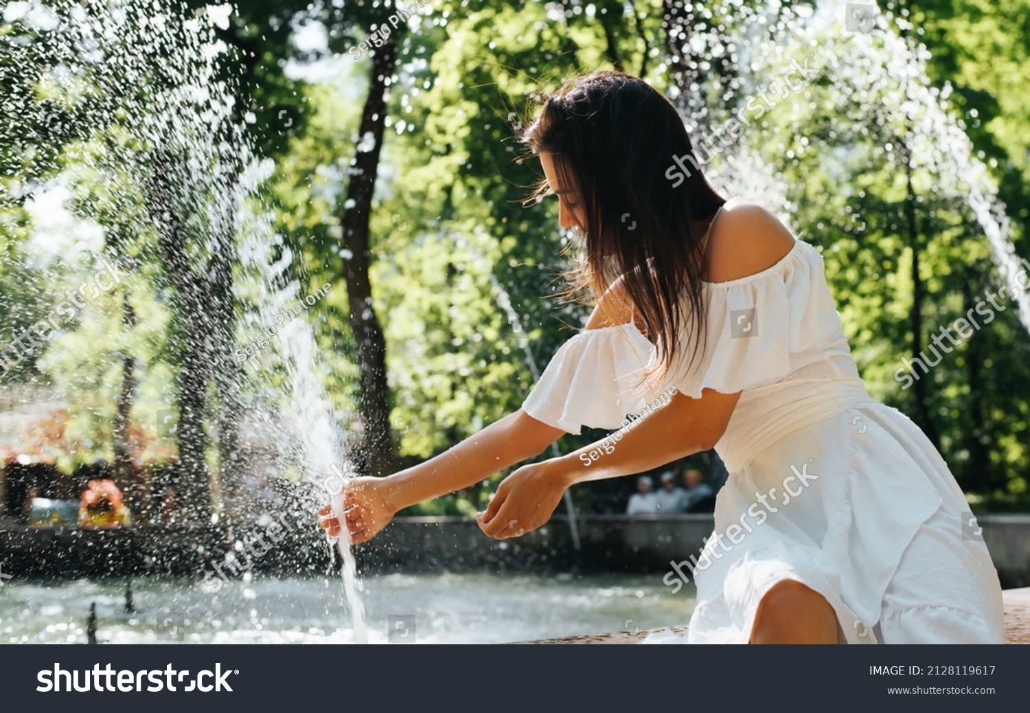 Young mixed race woman in white summer dress sitting by fountain on sunny day in park. Joyful positive woman cooling down by water on hot day outdoors. #2128119617