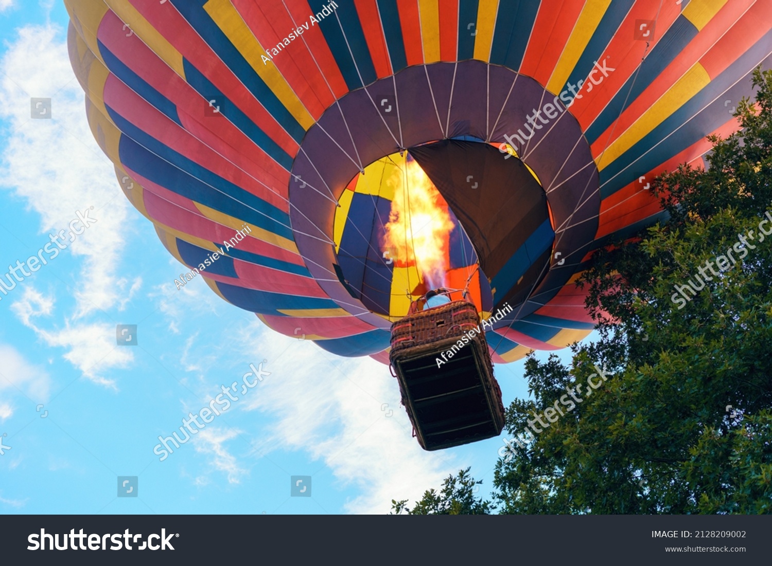 Colorful hot air balloon with people in a basket rises to the sky among the trees. #2128209002