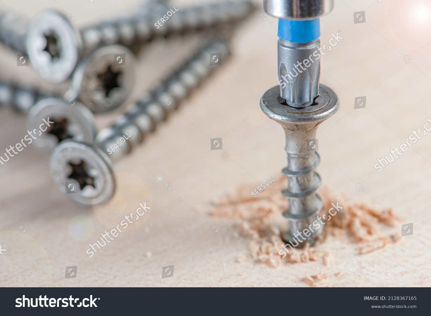 Screw the screw into the board. Furniture production, a screwdriver twists a self-tapping screw into a board. Several silver screws lie on the desktop #2128367165