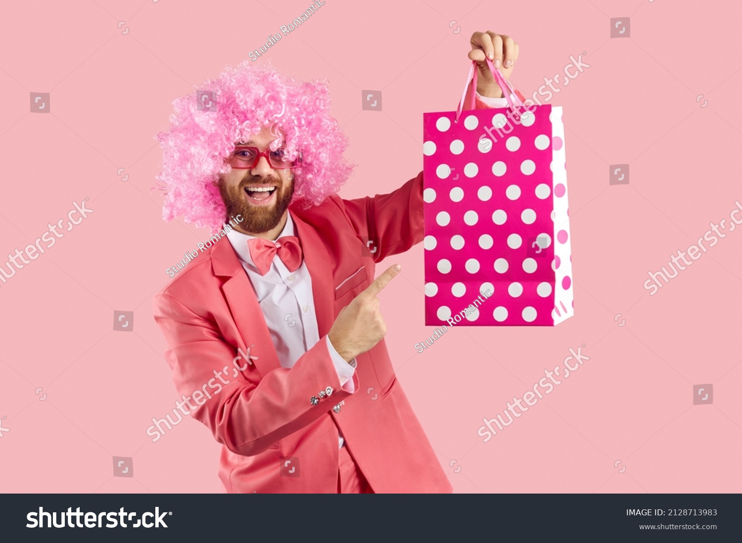 Funny crazy clown man in pink curly wig, sunglasses, funky suit and bow tie showing pink and white polka dot paper bag and smiling. Shopping, sale, presents, discounts, special offers concept #2128713983