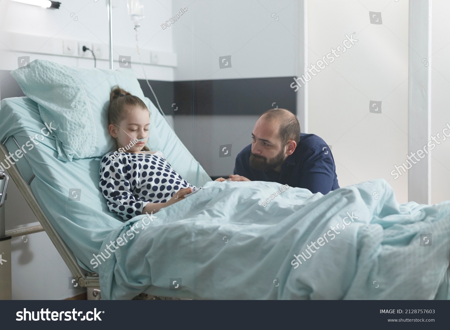 Worried sad uneasy father watching sick little daughter sleeping in hospital bed while in pediatric clinic patient room. Under treatment ill child resting while attentive parent taking care of her. #2128757603