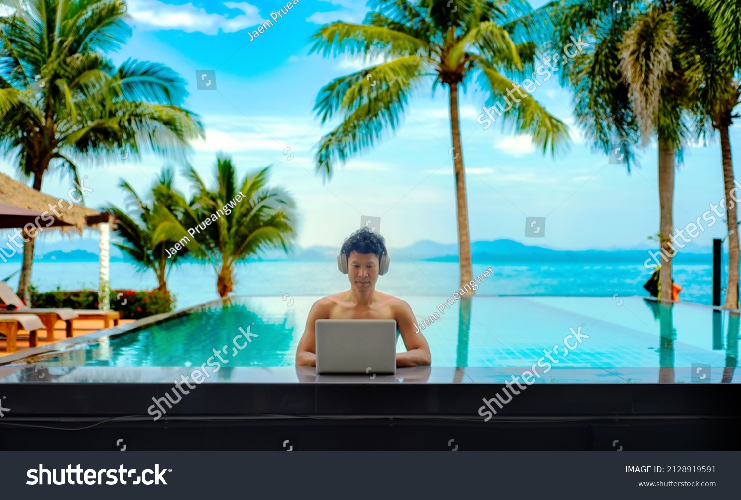An asian man working on his laptop at. a pool bar with his headphone on, behind him there is the view of the sea, coconut trees and the mountains #2128919591