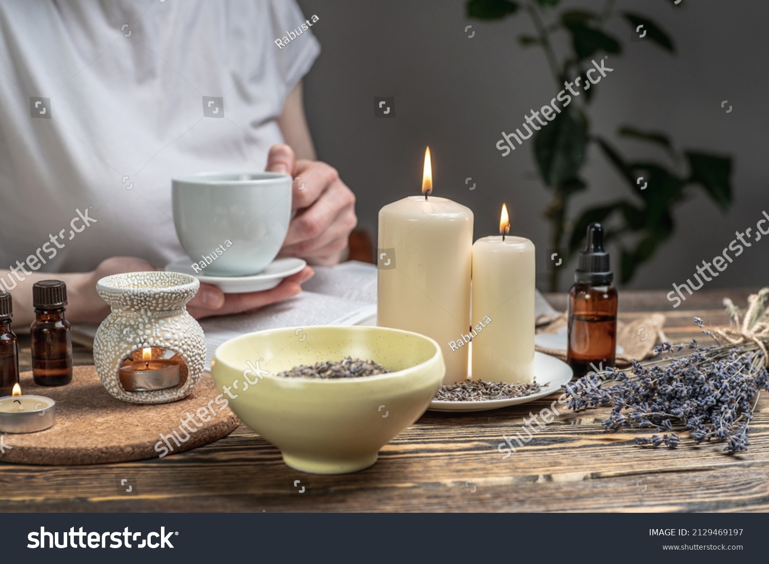 Woman is reading a book and drinking tea in atmosphere of harmony and relaxation. Aroma lamp with essential oils and burning candles on the wooden table. #2129469197