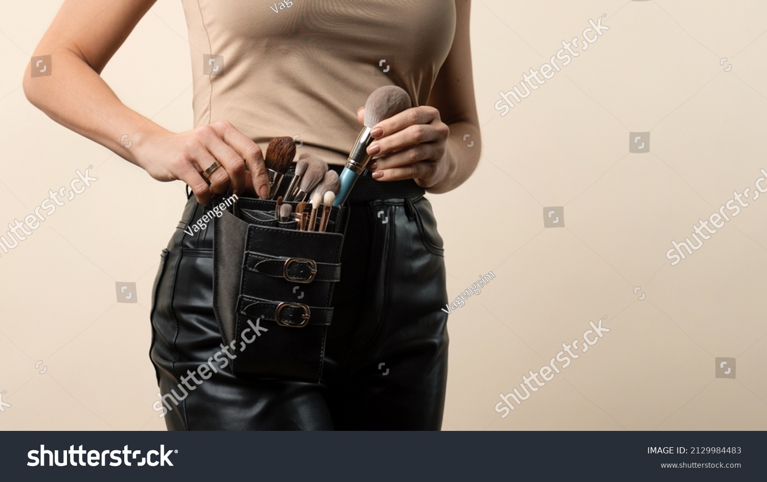 Thigh bag with different brushes on faceless professional makeup artist. Unrecognizable make-up artist with belt bag with tassels or makeup brushes over beige background #2129984483