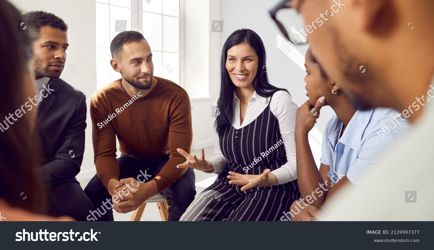 Female coach or team leader tells funny story or joke to diverse team during work meeting. Multiracial employees sitting in circle on chairs during informal brainstorming exchange ideas. #2129997377