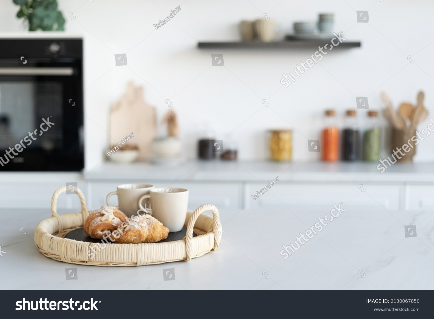Fresh croissants and cups of tea in a basket in contemporary kitchen interior. Kitchen appliances and decor on background. Homemade bakery concept. Modern white furniture #2130067850