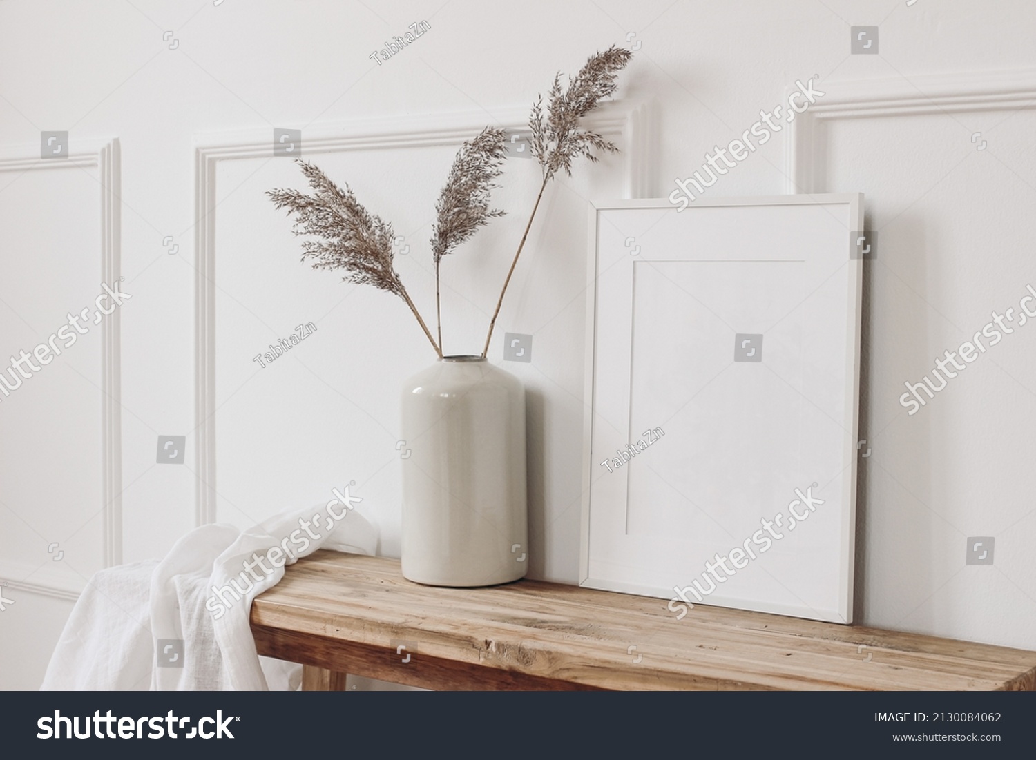 Elegant home interior decor still life photo. Vase with dry reed, grass on old wooden bench. Blank white picture frame mockup. Wall moulding background, trim decor. Empty copy space. Side view.  #2130084062