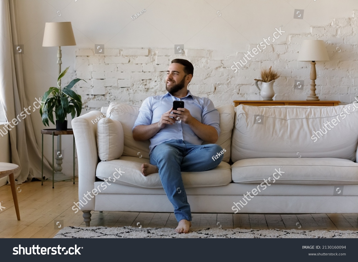 Smiling man sit on sofa at home with smartphone distracted from device look into distance spend weekend using modern cellphone, single guy enjoy e-dating services. Wireless tech, fun, leisure concept #2130160094